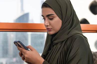 Tapping on a mobile phone screen, browsing social media on mobile, an Emirati Gulf Arab woman wearing a hijab with a green abaya sitting in an outdoor cafe, using modern technology devices