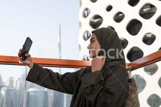 توثيق اللحظات التذكارية عبر جهاز الجوال,  قضاء الأوقات الممتعة في مقهى مرتفع مطل على المدينة, امرأة عربية خليجية اماراتية محجبة ترتدي عباءة خضراء وتحمل هاتفها