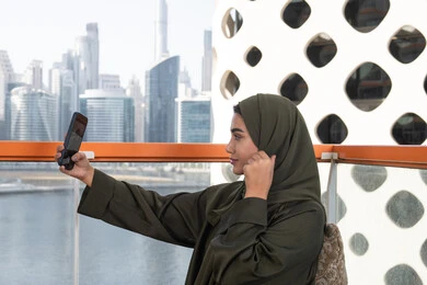 التقاط صور سيلفي تذكارية عبر الجوال, توثيق اللحظات السعيدة, قضاء الأوقات الممتعة في مقهى مرتفع مطل على المدينة, امرأة عربية خليجية اماراتية محجبة ترتدي عباءة خضراء وتحمل هاتفها