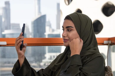 صورة مقربة لسيدة عربية خليجية اماراتية مبتسمة و ترتدي الحجاب مع عباءة خضراء  تلتقط صورة سيلفي للتذكار,  قضاء الأوقات الممتعة في مقهى مرتفع مطل على المدينة, مشاركة اللحظات السعيدة على وسائل التواصل الاجتماعي، استخدام الأجهزة التقنية الحديثة