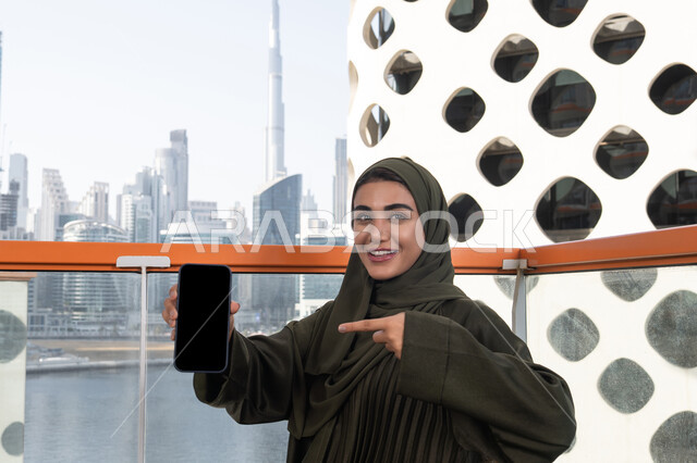 Pointing index finger at blank black phone screen, veiled Emirati Gulf Arab woman wearing green abaya sitting in outdoor cafe, gestures indicating happiness and pleasure, having fun
