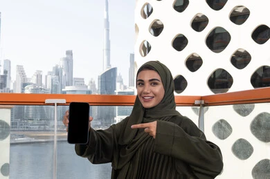 الإشارة بإصبع السبابة إلى شاشة الهاتف السوداء الفارغة, امرأة عربية خليجية اماراتية محجبة ترتدي عباءة خضراء تجلس في مقهى في الهواء الطلق, إيماءات تدل على السعادة و السرور، قضاء الأوقات الممتعة