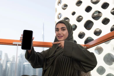 استخدام الهاتف الجوال والتقنيات الحديثة, قضاء الأوقات الممتعة في مقهى مرتفع مطل على المدينة, امرأة عربية خليجية اماراتية محجبة ترتدي عباءة خضراء مبتسمة تشير باصبع السبابة الى الهاتف المحمول و تقوم بعرض شاشة سوداء فارغة