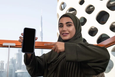 صورة مقربة لإمرأة عربية خليجية اماراتية محجبة ترتدي عباءة خضراء مبتسمة تشير باصبع السبابة الى الهاتف المحمول و تقوم بعرض شاشة سوداء فارغة, قضاء الأوقات الممتعة في مقهى مرتفع مطل على المدينة