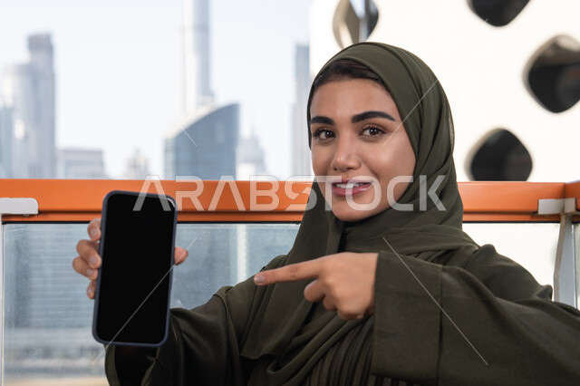 عرض شاشة سوداء فارغة عبر الهاتف المحمول, امرأة عربية خليجية اماراتية محجبة ترتدي عباءة خضراء مبتسمة تشير باصبع السبابة الى الجوال و تجلس في مقهى في الهواء الطلق