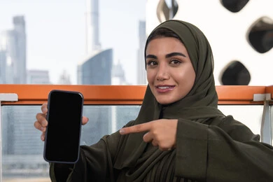 Blank black screen view on mobile phone, a veiled Arab Gulf Emirati woman wearing a green abaya smiling and pointing her index finger at the mobile phone and sitting in an outdoor cafe