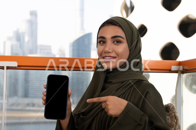 ايماءات وجه تدل على السعادة و السرور، قضاء الأوقات الممتعة في مقهى مرتفع مطل على المدينة, امرأة عربية خليجية اماراتية محجبة ترتدي عباءة خضراء مبتسمة تشير باصبع السبابة الى الهاتف المحمول و تقوم بعرض شاشة سوداء فارغة