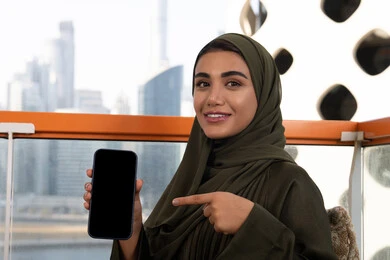 Facial expressions indicating happiness and pleasure, having a good time in a high-rise cafe overlooking the city, a veiled Emirati Gulf Arab woman wearing a green abaya, smiling, pointing her index finger at a mobile phone displaying a blank black screen