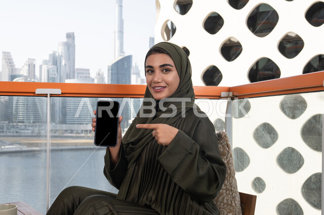 Having fun in a high-rise cafe overlooking the city, a veiled Emirati Gulf Arab woman wearing a green abaya, smiling and pointing her index finger at her mobile phone displaying a blank black screen
