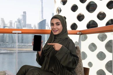 قضاء الأوقات الممتعة في مقهى مرتفع مطل على المدينة, امرأة عربية خليجية اماراتية محجبة ترتدي عباءة خضراء مبتسمة تشير باصبع السبابة الى الهاتف المحمول و تقوم بعرض شاشة سوداء فارغة