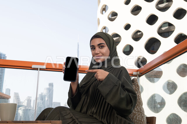 تطبيقات الهواتف الذكية، عرض شاشة سوداء فارغة عبر الهاتف المحمول, امرأة عربية خليجية اماراتية محجبة ترتدي عباءة خضراء مبتسمة تشير باصبع السبابة الى الجوال و تجلس في مقهى في الهواء الطلق