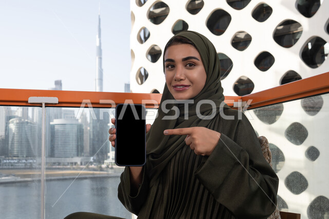 شاشة هاتف محمول سوداء فارغة, قضاء الأوقات الممتعة في مقهى مرتفع مطل على المدينة, امرأة عربية خليجية اماراتية محجبة ترتدي عباءة خضراء مبتسمة تشير باصبع السبابة الى الجوال