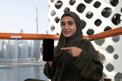 Blank black mobile phone screen, having fun in a high-rise cafe overlooking the city, a veiled Emirati Gulf Arab woman wearing a green abaya, smiling, pointing her index finger at the mobile phone