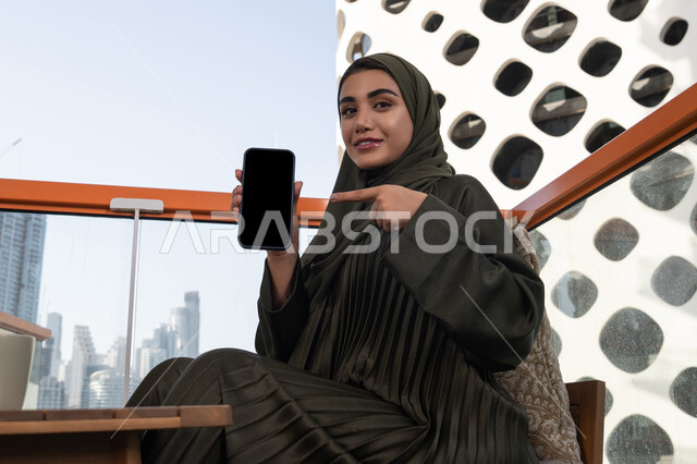 النظر الى الكاميرا بايماءات السرور والسعادة، امرأة عربية خليجية اماراتية محجبة ترتدي عباءة خضراء مبتسمة تشير باصبع السبابة الى الهاتف المحمول, الجلوس في مقهى مرتفع مطل على المدينة