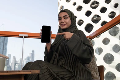 Looking at the camera with gestures of pleasure and happiness, a veiled Emirati Gulf Arab woman wearing a green abaya, smiling and pointing her index finger at her mobile phone, sitting in a high cafe overlooking the city