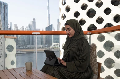 Typing and tapping on a tablet, a young Arab Gulf Emirati woman wearing a green abaya and a hijab, smiling, working on a tablet in a cafe, using modern and advanced technologies
