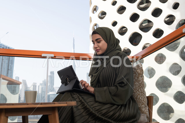 استعمال التكنولوجيا في العمل عن بعد، شابة عربية خليجية اماراتية محجبة ترتدي عباءة خضراء مبتسمة تعمل على التابلت بجانبها مشروب ساخن, تصفح الانترنت واستخدام تقنيات حديثة