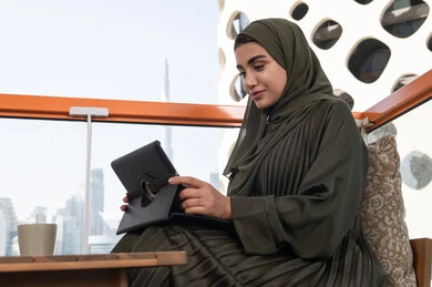 Taking advantage of modern techniques and advanced technology at work, a young veiled Emirati Gulf Arab woman wearing a green abaya, smiling, working on a tablet with a hot drink next to her, using a tablet in a modern cafe overlooking the city