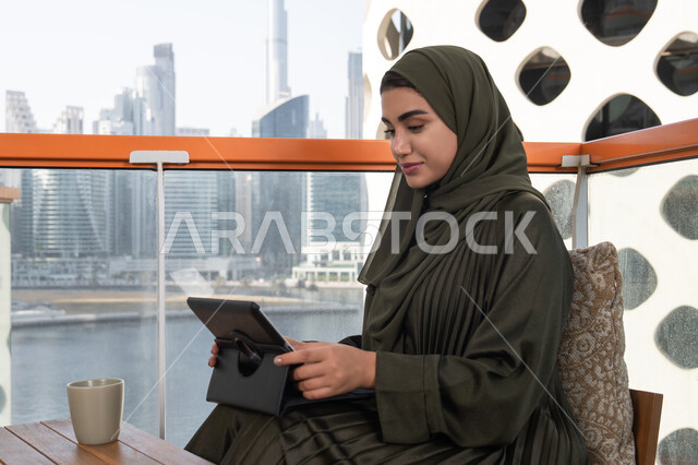 Modern cafe overlooking the city, a young veiled Emirati Gulf Arab woman wearing a green abaya, smiling, working on a tablet with a hot drink next to her, working remotely via the internet