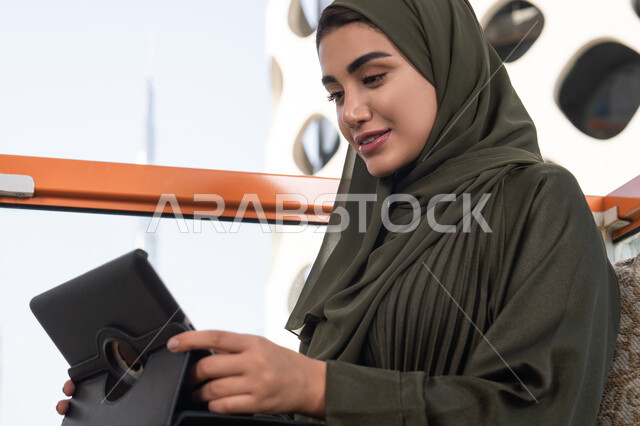 Completing work tasks remotely through a tablet, an Emirati Gulf Arab woman wearing a hijab with a green abaya sitting in an outdoor cafe, chatting and talking via video calls, gestures of happiness and pleasure