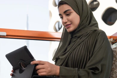 إنجاز مهام العمل عن بعد من خلال جهاز التابلت, سيدة عربية خليجية اماراتية ترتدي الحجاب مع عباءة خضراء  تجلس في مقهى في الهواء الطلق, الدردشات والمحادثات عبر مكالمات الفيديو, ايماءات السعادة والسرور