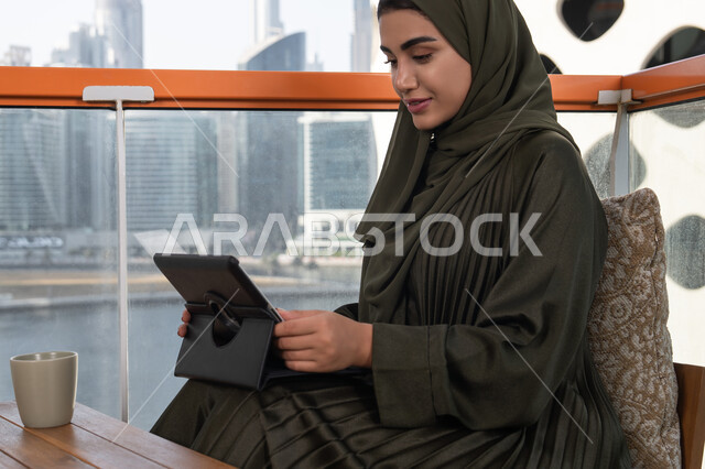 استخدام الجهاز اللوحي في مقهى عصري بإطلالة على المدينة, امرأة عربية خليجية اماراتية مبتسمة محجبة ترتدي عباءة خضراء بجانبها مشروب ساخن, العمل عن بعد من خلال الانترنت