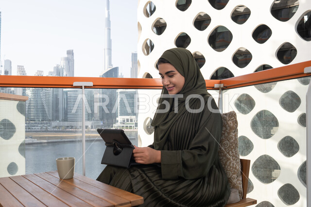 Using modern and advanced technology, making video calls via tablet, a young veiled Emirati Gulf Arab woman wearing a green abaya and hijab, smiling, working on a tablet in a cafe, completing work tasks remotely via the internet