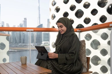 استخدام التكنولوجيا الحديثة والمتطورة، إجراء مكالمات فيديو عبر الجهاز اللوحي، شابة عربية خليجية اماراتية محجبة ترتدي عباءة خضراء وحجاب مبتسمة  تعمل على التابلت في احدى المقاهي الراقية، إنجاز مهام العمل عن بعد من خلال الانترنت