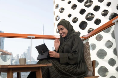 Raising hand with greeting expression, a smiling veiled Saudi Arabian Gulf woman using a tablet to make a video call, sitting in an upscale cafe, completing work tasks remotely