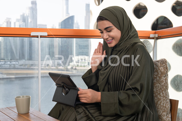 إلقاء التحية والترحيب والسلام, ايماءات السرور والسعادة, العمل عن بعد عن طريق جهاز التابلت, إمرأة عربية اماراتية خليجية محجبة ترتدي حجاب و عباءة خضراء تجلس في مقهى مرتفع مطل على المدينة, بجانبها مشروب ساخن