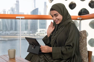Greeting, welcoming and peace, gestures of pleasure and happiness, working remotely via tablet, a veiled Emirati Gulf Arab woman wearing a green abaya and hijab sitting in a high cafe overlooking the city, next to her is a hot drink