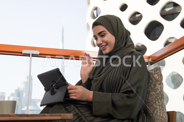 الدردشة و التواصل الاجتماعي, إجراء مكالمة فيديو عبر التابلت, ايماءات تدل على التحية والسلام, امرأة عربية خليجية اماراتية ترتدي العباءة والحجاب تجلس في مقهى هادئ, تقنيات العمل عن بعد