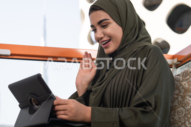 تصفح الانترنت واستخدام تقنيات حديثة, رفع الكف للأعلى بايماءات التحية والسلام,  إجراء مكالمة فيديو عبر جهاز التابلت, ايماءات السرور والسعادة, العمل عن بعد من خلال الانترنت, إمرأة عربية اماراتية خليجية محجبة  ترتدي حجاب و عباءة خضراء تجلس في مقهى مرتفع مطل على المدينة