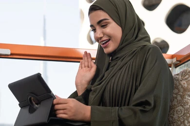 Browsing the internet and using modern technologies, raising the palm up in greeting and peace gestures, making a video call via a tablet, gestures of pleasure and happiness, working remotely via the internet, a veiled Emirati Gulf Arab woman wearing a green abaya and hijab sitting in a high cafe overlooking the city