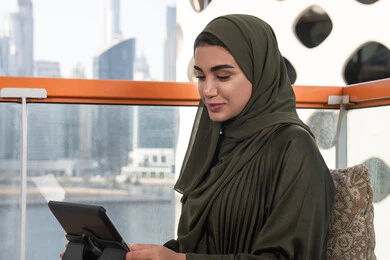 امرأة عربية خليجية اماراتية مبتسمة محجبة ترتدي عباءة خضراء تستخدم التابلت في مقهى مرتفع مطل على المدينة ,انجاز العمل عن بعد في الهواء الطلق