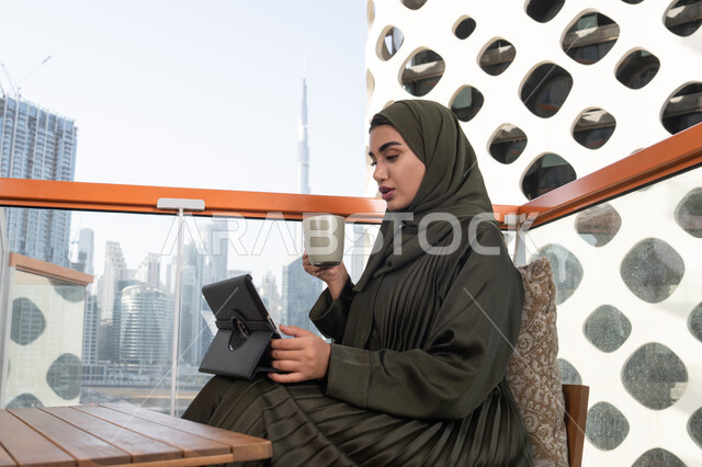 تعابير وايماءات الاندماج, امرأة عربية خليجية اماراتية محجبة ترتدي عباءة خضراء وتجلس في مقهى فاخر,الاستمتاع بمشروب ساخن, العمل عن بعد عن طريق التابلت