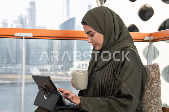 مفهوم إنجاز المشاريع والمهام العمل عن بعد، تصفح مواقع التواصل الاجتماعي، التواصل مع الأهل والأصدقاء عبر الايباد, امرأة عربية خليجية اماراتية محجبة ترتدي عباءة خضراء وتجلس في مقهى فاخر,الاستمتاع بمشروب ساخن