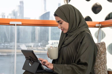 مفهوم إنجاز المشاريع والمهام العمل عن بعد، تصفح مواقع التواصل الاجتماعي، التواصل مع الأهل والأصدقاء عبر الايباد, امرأة عربية خليجية اماراتية محجبة ترتدي عباءة خضراء وتجلس في مقهى فاخر,الاستمتاع بمشروب ساخن