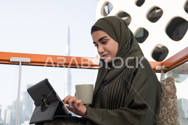 بيئة عمل مريحة في احدى المقاهي الراقية, شابة عربية خليجية اماراتية محجبة ترتدي عباءة و حجاب و تحمل كوب من المشروب الساخن, تصفح الانترنت واستخدام تقنيات حديثة