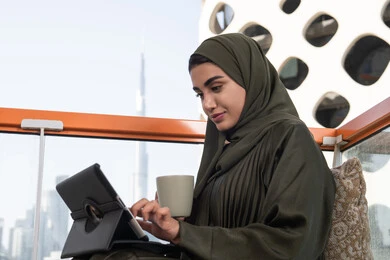 بيئة عمل مريحة في احدى المقاهي الراقية, شابة عربية خليجية اماراتية محجبة ترتدي عباءة و حجاب و تحمل كوب من المشروب الساخن, تصفح الانترنت واستخدام تقنيات حديثة