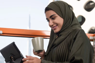 اجراء مقابلات عمل اونلاين عبر التابلت،سيدة عربية خليجية سعودية مرتدية عباءة و حجاب, الدردشات والمحادثات عبر مكالمات الفيديو, ايماءات السعادة والسرور, الاستمتاع بكوب من المشروب الساخن