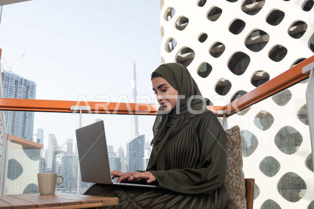 الكتابة والنقر على الحاسوب محمول، شابة عربية خليجية اماراتية محجبة ترتدي عباءة خضراء وحجاب مبتسمة  تعمل على اللابتوب  في احدى المقاهي، استخدام التقنيات الحديثة والمتطورة, إنجاز مهام العمل عن بعد من خلال الانترنت