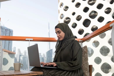 الكتابة والنقر على الحاسوب محمول، شابة عربية خليجية اماراتية محجبة ترتدي عباءة خضراء وحجاب مبتسمة  تعمل على اللابتوب  في احدى المقاهي، استخدام التقنيات الحديثة والمتطورة, إنجاز مهام العمل عن بعد من خلال الانترنت