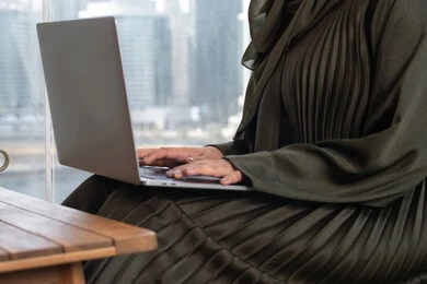 متابعة العمل عن بعد عبر الحاسوب المحمول, صورة مقربة لامرأة عربية خليجية اماراتية  تعمل على اللابتوب في احدى المقاهي الفارهة و ترتدي عباءة خضراء,  الكتابة والنقر على اللابتوب, تصفح الانترنت واستخدام التقنيات الحديثة