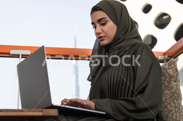 استخدام الأجهزة والتقنيات الحديثة والمتطورة، شابة عربية إماراتية خليجية محجبة ترتدي عباءة خضراء في مقهى مرتفع مطل على المدينة, العمل على جهاز الكمبيوتر المحمول