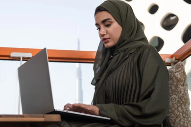 استخدام الأجهزة والتقنيات الحديثة والمتطورة، شابة عربية إماراتية خليجية محجبة ترتدي عباءة خضراء في مقهى مرتفع مطل على المدينة, العمل على جهاز الكمبيوتر المحمول