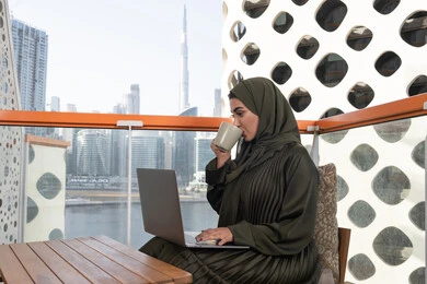 احتساء مشروب ساخن, شابة عربية خليجية اماراتية تجلس في مقهى راقي مطل على المدينة ترتدي الحجاب وعباءة خضراء , استخدام جهاز الحاسوب المحمول والعمل عن بعد من خلال الانترنت