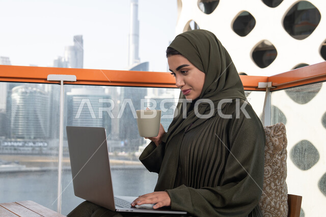 Accomplishing tasks in a calm and comfortable atmosphere, enjoying a hot drink, a veiled Arab Gulf Emirati woman wearing a green abaya and hijab, using a laptop to work remotely