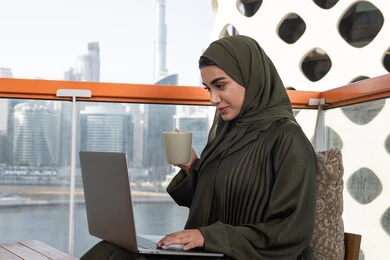 إنجاز المهام في جو هادئ ومريح, الاستمتاع بتناول مشروب ساخن, سيدة محجبة عربية خليجية اماراتية ترتدي عباءة خضراء و حجاب, استخدام الكمبيوتر المحمول للعمل عن بعد