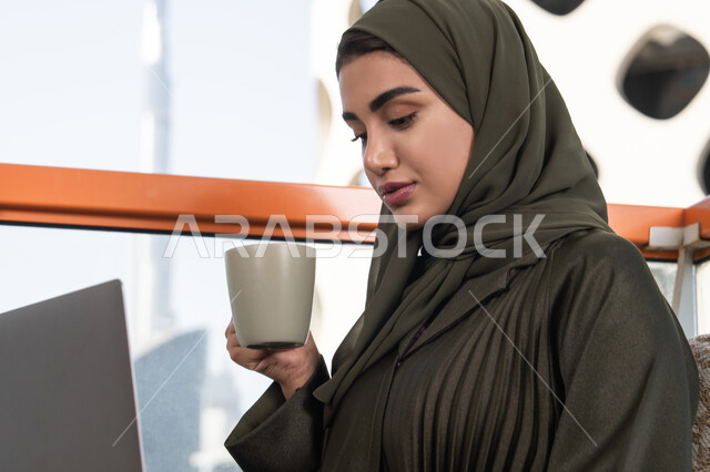 الاستغراق في العمل, الاستمتاع بكوب من المشروب الساخن, شابة عربية خليجية اماراتية محجبة ترتدي عباءة خضراء و حجاب, العمل عن بعد باستخدام الانترنت عبر الحاسوب المحمول في مقهى مرتفع في الهواء الطلق